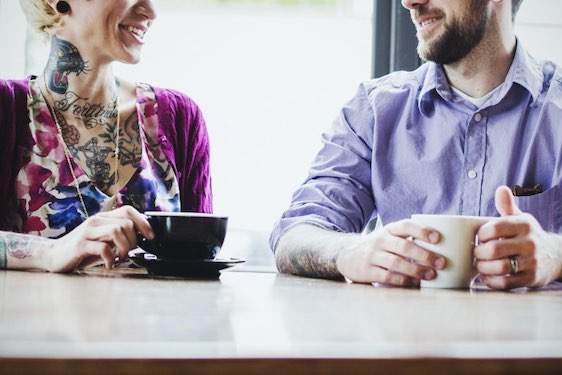 A trendy hipster man and woman with tattoos smile and talk as they enjoy a coffee and latte at a bright industrial styled cafe setting, sitting at a large bamboo wood table. Horizontal with copy space.