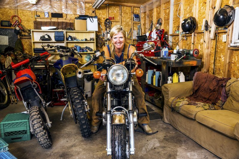 A pretty middle-aged woman in her garage full of motorcycles, dirt bikes and tools.  She is sitting astride a vintage motorcycle while smiling and looking at the camera and making eye contact. An authentic scene with a real person.