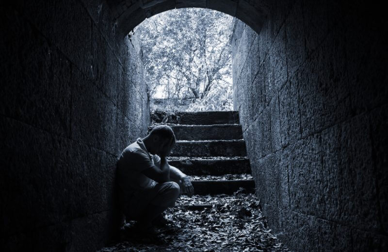 Young sad man sitting inside of dark stone tunnel, blue tonal correction photo filter effect