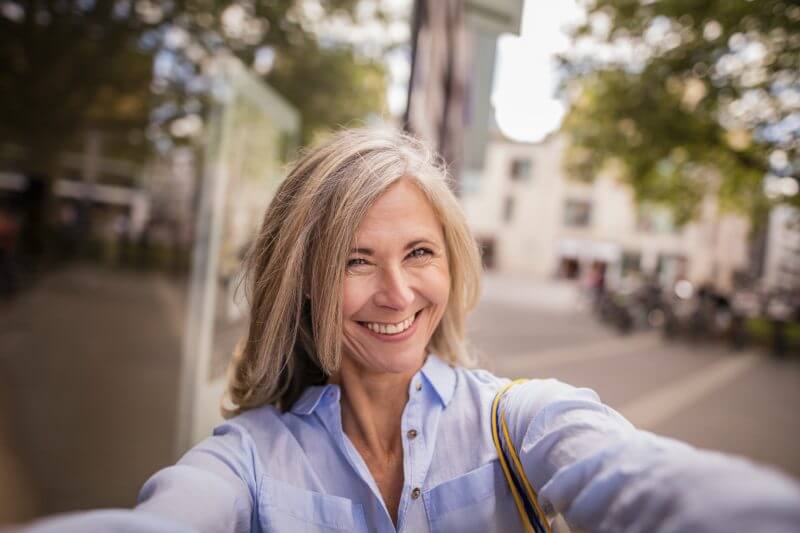 Beautiful smiling mature woman with grey hair taking a selfie while standing on a city street