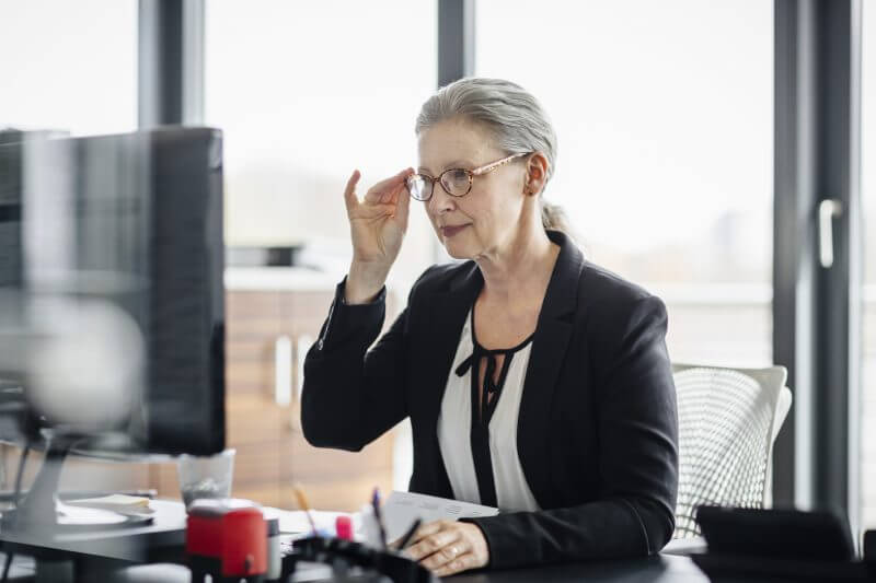 Portrait of a senior businesswoman sitting on her desk in a modern office and. She's working on a computer and wearing glasses and has silver hair.