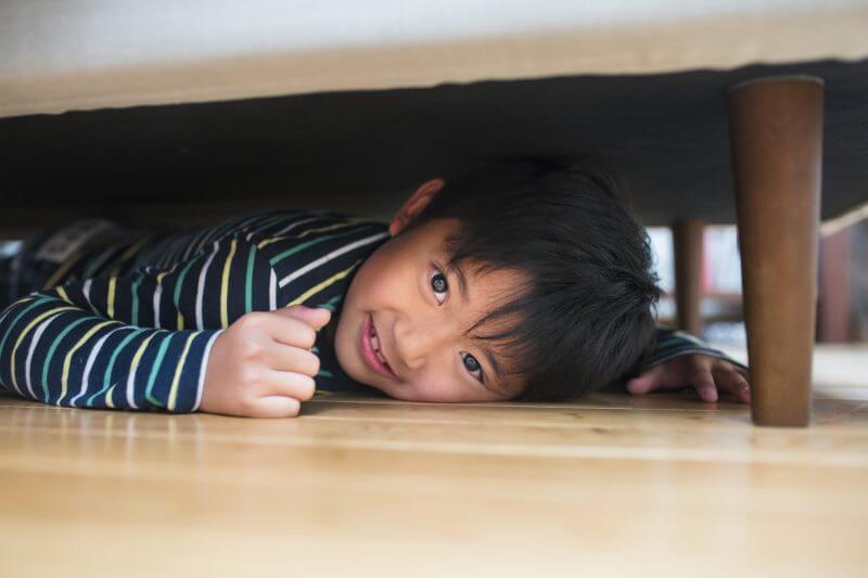 Young boy with a mischievous smile on his face hiding under the sofa. Kyoto, Japan. May 2016.