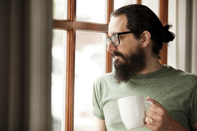A man with a beard, man bun and hipster glasses holding a cup of coffee as he gazes though a window lost in thought.