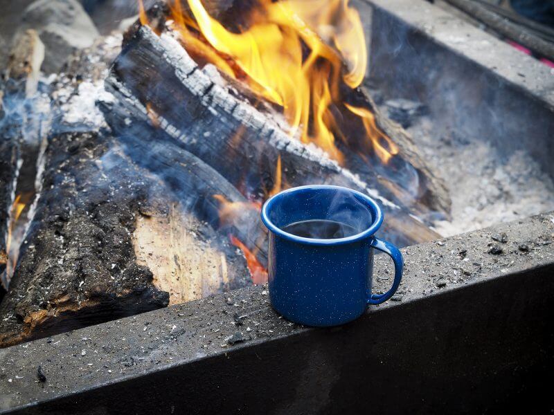 Steaming cup of camp coffee staying warm on edge of campfire.