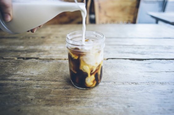 A female hand pours cream milk into a mason jar of cold brew coffee.